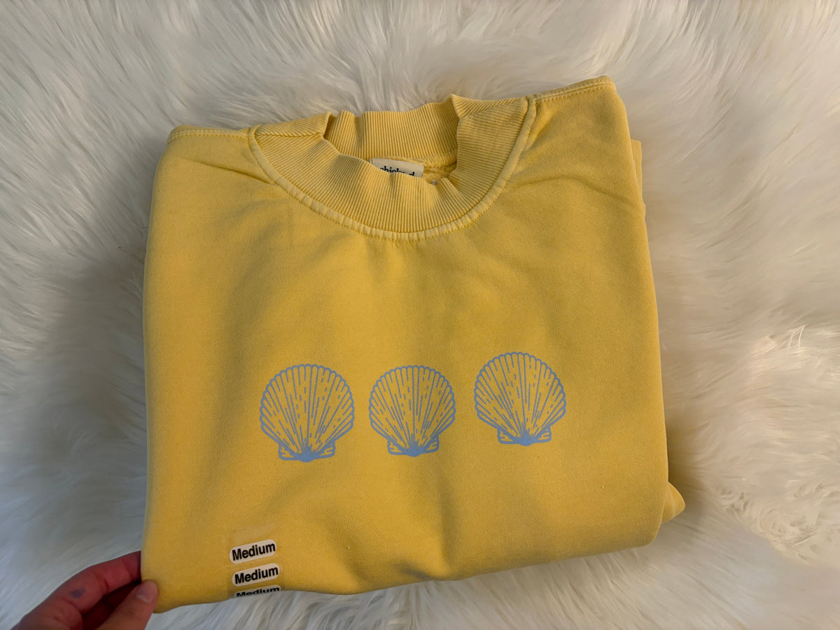 A folded yellow sweatshirt on a white faux fur background, featuring three light blue scallop shell graphics on the front and a small black-and-white size label that reads Medium.